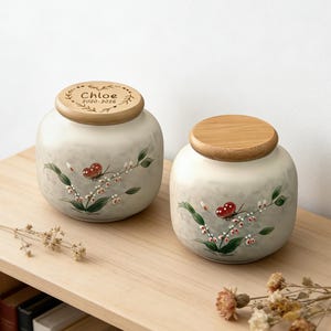 Puede incluir: Dos urnas de cerámica con tapas de madera. Las urnas son de color blanquecino con un diseño floral y de mariposas. Una tapa tiene el nombre "Chloe" y los años "2020-2026" grabados. Las urnas están sobre una superficie de madera.