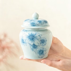 May include: A small, light blue ceramic jar with a lid, decorated with blue floral patterns. The jar is held in open hands, showcasing its delicate design. The jar has a rounded shape and a small knob on the lid.