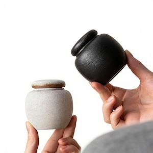 Puede incluir: Dos pequeños tarros de cerámica. Uno es blanco moteado con tapa marrón, y el otro es negro mate. Los tarros se sostienen sobre un fondo blanco. Los tarros tienen forma redondeada y superficie texturizada.