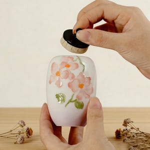 May include: A white ceramic container with a floral design in pink and orange. A small, round, black-topped brush is held above the container. The container has a rounded rectangular shape and is decorated with pink flowers and green leaves.