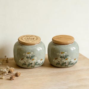 May include: Two ceramic jars with wooden lids. The jars are light green with a crackle glaze and painted white daisy flowers. One lid is engraved with the name "Chloe" and the years "2020-2026".