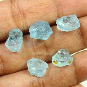 Pode incluir: Cinco pedras brutas de aguamarina azul clara. As pedras variam em tamanho e forma, com uma qualidade translúcida. As aguamarinas são exibidas em um fundo cor de pele, mostrando sua cor e textura naturais.