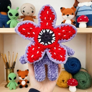 May include: A handmade, plush Demogorgon from the series, Stranger Things, in red, purple, and white. The flower-like creature has a large, open mouth with white teeth. Other crocheted toys, including aliens, foxes, and mushrooms, are visible in the background.