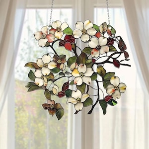 May include: Stained glass window hanging featuring a dogwood branch with white and pink flowers, green leaves, and red accents. The artwork is suspended by a chain and hangs in front of a window with white curtains.