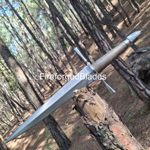 May include: A long, silver sword with a textured brown handle and a crossguard. The sword is resting on a piece of wood in a forest setting. The text "FireforgedBlades" is visible on the image.