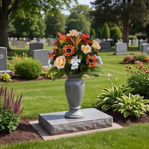 May include: A gray stone vase sits on a rectangular granite base, filled with an arrangement of artificial flowers including orange sunflowers, cream-colored roses, and small white blossoms, accented with green foliage and a small red pumpkin.
