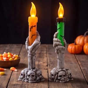 May include: Two battery-operated LED taper candles held by skeletal hand candle holders. One candle is orange with black bat silhouettes, the other is green with black bat silhouettes. Both bases feature a cluster of detailed human skulls.