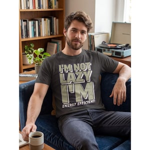 Puede incluir: Camiseta gris oscuro con el texto "I'M NOT LAZY I'M ENERGY EFFICIENT" en letras blancas y verde claro. La camiseta la lleva una persona sentada en un sofá azul. Una taza de café se sostiene en la mano de la persona.