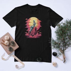 May include: Black t-shirt with a graphic design of a skeleton seated on a rock formation, set against a sunset. The design features a yellow sun, pink clouds, and a seated skeleton figure.
