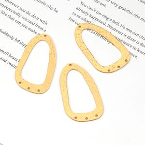 May include: Three gold-colored, open-frame pendants with a hammered texture. Each pendant has an elongated, asymmetrical shape with multiple small holes for attaching to other components. The pendants are lying on a white surface.