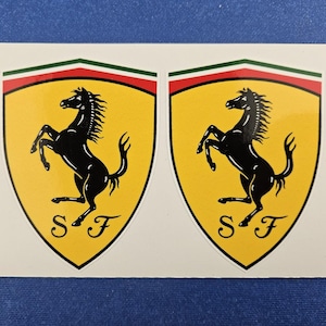 May include: Two stickers featuring the Ferrari logo. Each sticker has a yellow shield shape with a black prancing horse, the letters "S" and "F" at the bottom, and a green, white, and red stripe at the top.