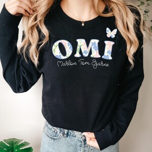 May include: Black sweatshirt with the word "OMI" in a floral design, with the names "Marlon Tom Bjarne" below. A small butterfly graphic is on the upper right. The sweatshirt is a casual style.
