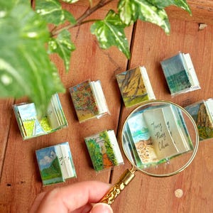 May include: Miniature books with landscape paintings are arranged on a wooden surface, some are open. A magnifying glass is held over one book, revealing the text "Tung Ping Chan". Green and white leaves are in the background.