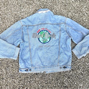 May include: A light blue denim jacket featuring a back graphic with the text Hard Rock Cafe, Save The Planet, and Las Vegas. The design includes a globe illustration in blue and green colors, with silver lettering for the brand name.