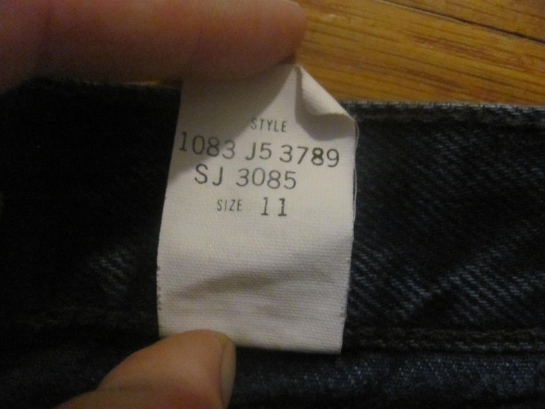 May include: A white tag with black text that reads "Style 1083 J5 3789 SJ 3085 Size 11" attached to a dark blue denim fabric.