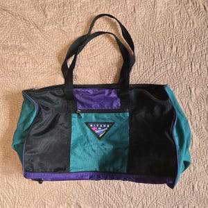 May include: A rectangular duffel bag with black handles and a zippered top. The bag features panels of black, teal, and purple fabric. A small teal pocket with a triangular logo is on the front. The bag is designed for travel or carrying items.