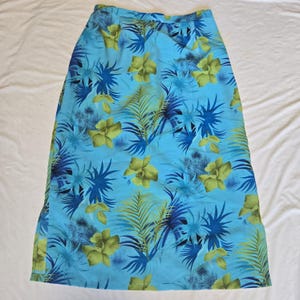 May include: A long, light blue skirt with a tropical floral pattern. The skirt features a repeating design of yellow-green flowers, blue spiky leaves, and green palm fronds. The skirt is made of a lightweight fabric.