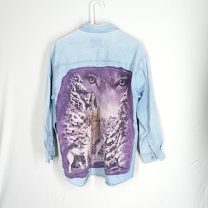 May include: A light blue denim shirt with a purple and white graphic print of a Native American woman standing with two wolves in a snowy forest. The graphic features the eyes of a wolf looking out from the top of the image.