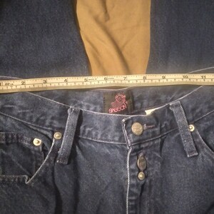 May include: A close-up of a pair of dark blue denim jeans with a Grecon label. The jeans have a button fly and five pockets. A measuring tape is placed across the waistband, showing a measurement of 30 inches.