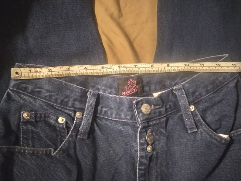 May include: A close-up of a pair of dark blue denim jeans with a Grecon label. The jeans have a button fly and five pockets. A measuring tape is placed across the waistband, showing a measurement of 30 inches.