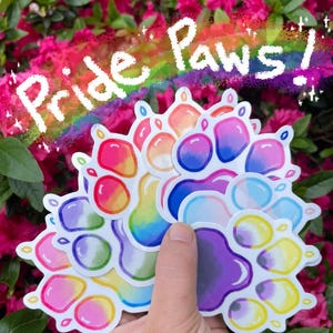 May include: A collection of colorful paw print stickers with a rainbow design. The stickers have a gradient of colors, including pink, orange, yellow, green, blue, and purple. The text "Pride Paws!" is written above the stickers.