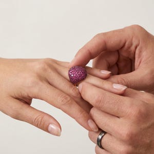 Op de afbeelding: Een gouden ring met een grote, ronde cluster van kleine, dieproze edelstenen. De ring wordt door een andere persoon om de vinger van een persoon gedaan. De achtergrond is effen licht van kleur.