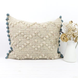 May include: A cream-colored crocheted pillow with a textured, bumpy surface. The pillow has a blue and white trim with pom-poms along the edges.