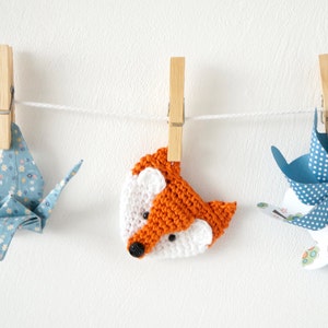 May include: Three handmade decorations hanging on a white string with wooden clothespins. The decorations are a blue origami crane, an orange crocheted fox, and a blue paper pinwheel with white polka dots.