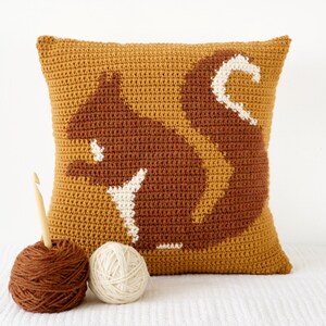 May include: A crocheted pillow with a brown squirrel design on a mustard yellow background. The pillow is sitting on a white surface with two balls of yarn and a crochet hook.