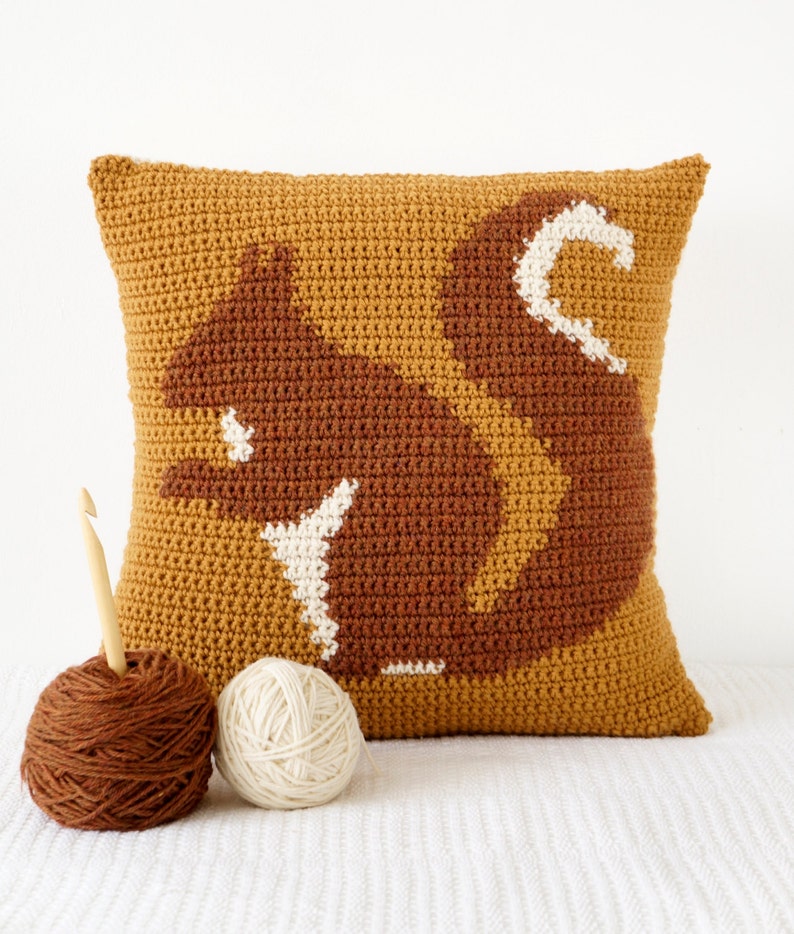 K&ouml;nnte beinhalten: Ein geh&auml;keltes Kissen mit einem braunen Eichh&ouml;rnchen-Design auf einem senfgelben Hintergrund. Das Kissen liegt auf einer wei&szlig;en Oberfl&auml;che mit zwei Wollkn&auml;ueln und einer H&auml;kelnadel.