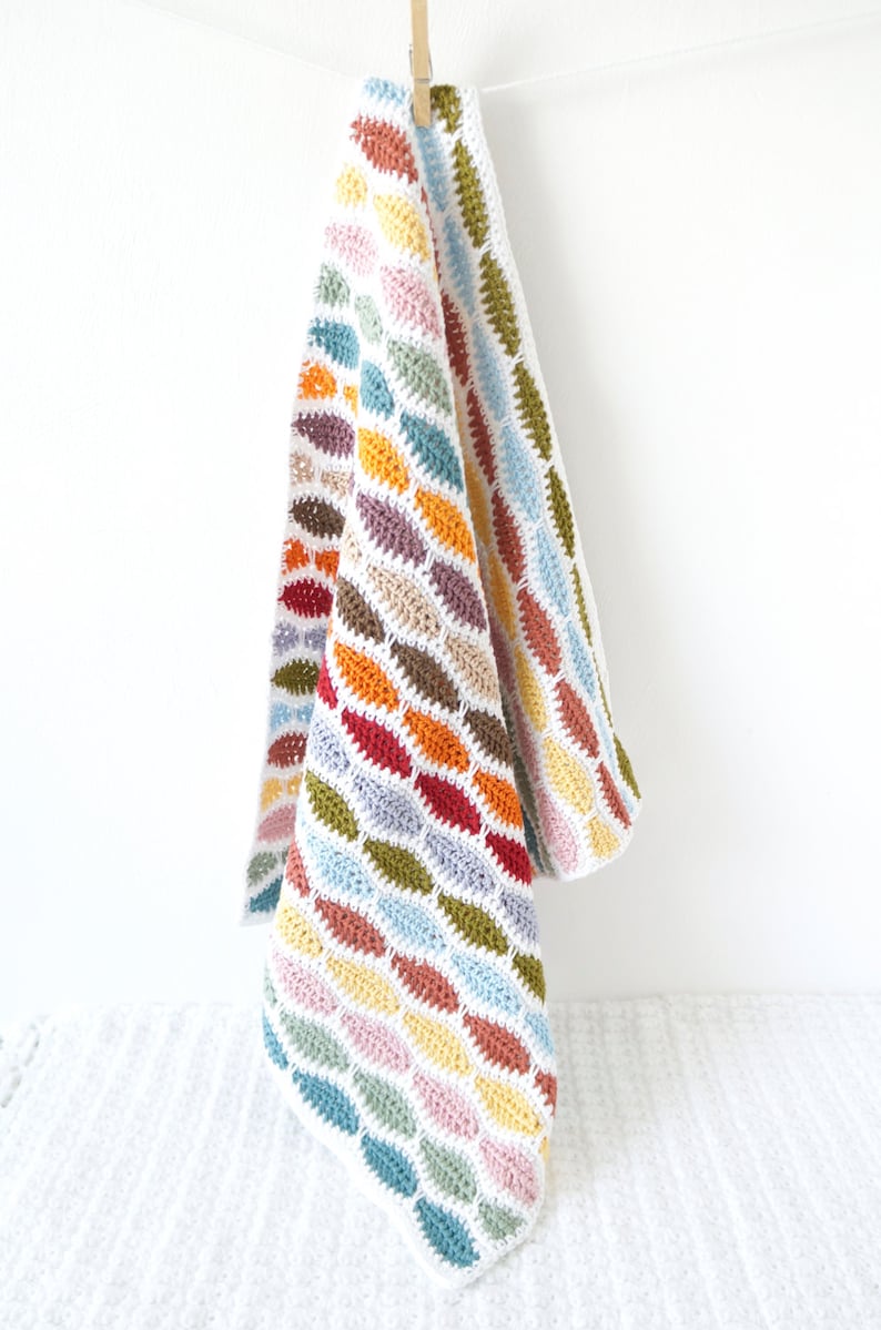 May include: A colorful, handmade crochet blanket with a repeating diamond pattern in various colors, including red, orange, yellow, green, blue, and purple. The blanket is hanging from a clothesline with a wooden clothespin.