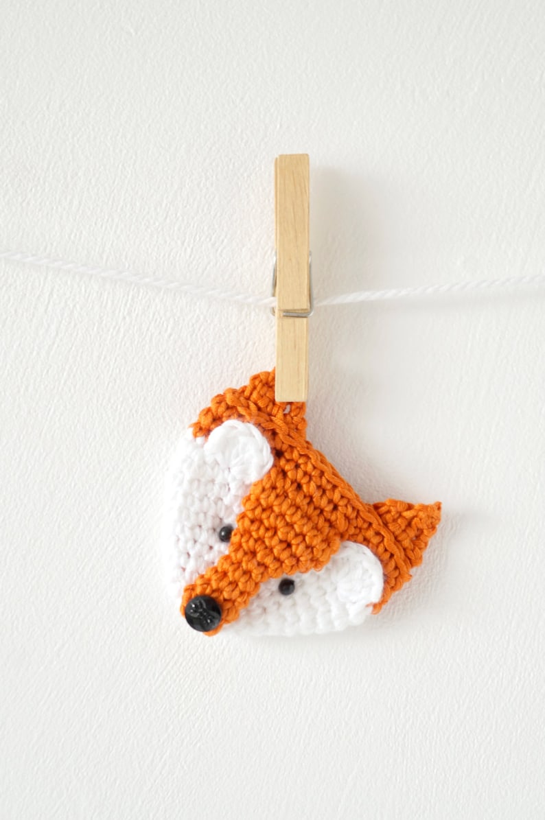 May include: A crocheted fox head ornament in orange and white with black button eyes, hanging from a wooden clothespin on a white string.