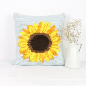 Puede incluir: Una almohada de crochet con un dise&ntilde;o de girasol. El girasol es amarillo y naranja con un centro marr&oacute;n. La almohada es azul claro.