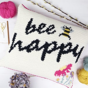 May include: Cream crochet cushion featuring the words "bee happy" in black script, a bee graphic, and floral details. The cushion has a pink border. Craft supplies, including yarn and scissors, are visible in the image.