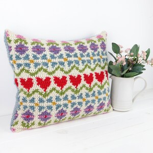 Op de afbeelding: Een gehaakt kussen met een kleurrijk geometrisch patroon met harten, bloemen en golvende lijnen in tinten rood, roze, groen, blauw en geel.