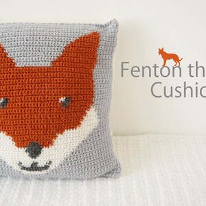 May include: A gray crocheted pillow with an orange fox face design. The fox has white fur around its mouth and black eyes and nose. The text "Fenton the Fox Cushion" is in the upper right corner of the image.