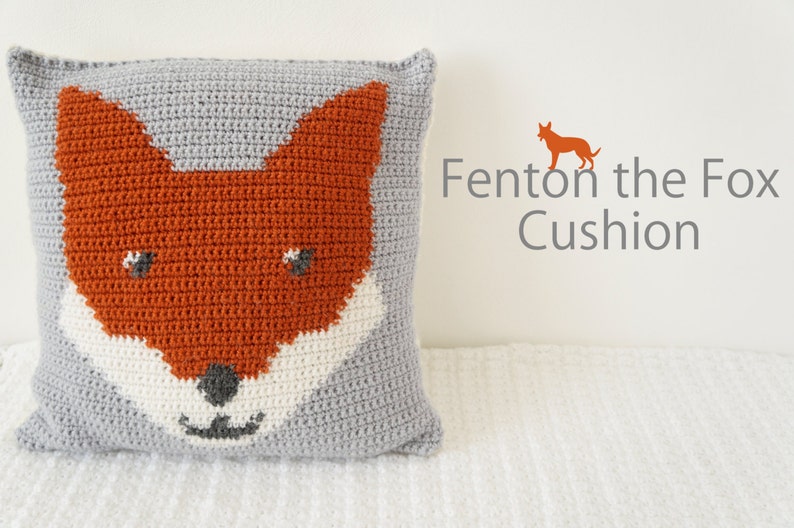 May include: A gray crocheted pillow with an orange fox face design. The fox has white fur around its mouth and black eyes and nose. The text "Fenton the Fox Cushion" is in the upper right corner of the image.