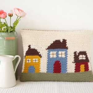 May include: A crocheted pillow with a design of three houses. The houses are yellow, blue, and red with brown roofs. The pillow is on a white surface.