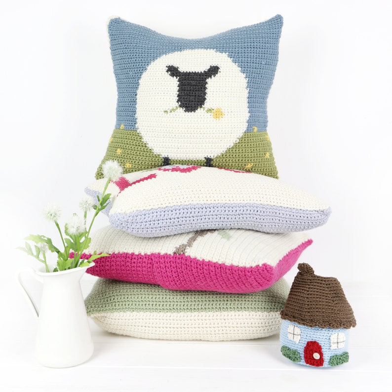 Puede incluir: Una pila de cuatro almohadas de crochet con diferentes colores y dise&ntilde;os. La almohada superior presenta una oveja blanca con ojos negros y una flor amarilla en la boca. La segunda almohada es blanca con un dise&ntilde;o de p&aacute;jaro rojo. La tercera almohada es de color morado claro y la almohada inferior es verde. Una peque&ntilde;a casa de crochet marr&oacute;n con una puerta roja est&aacute; en primer plano.
