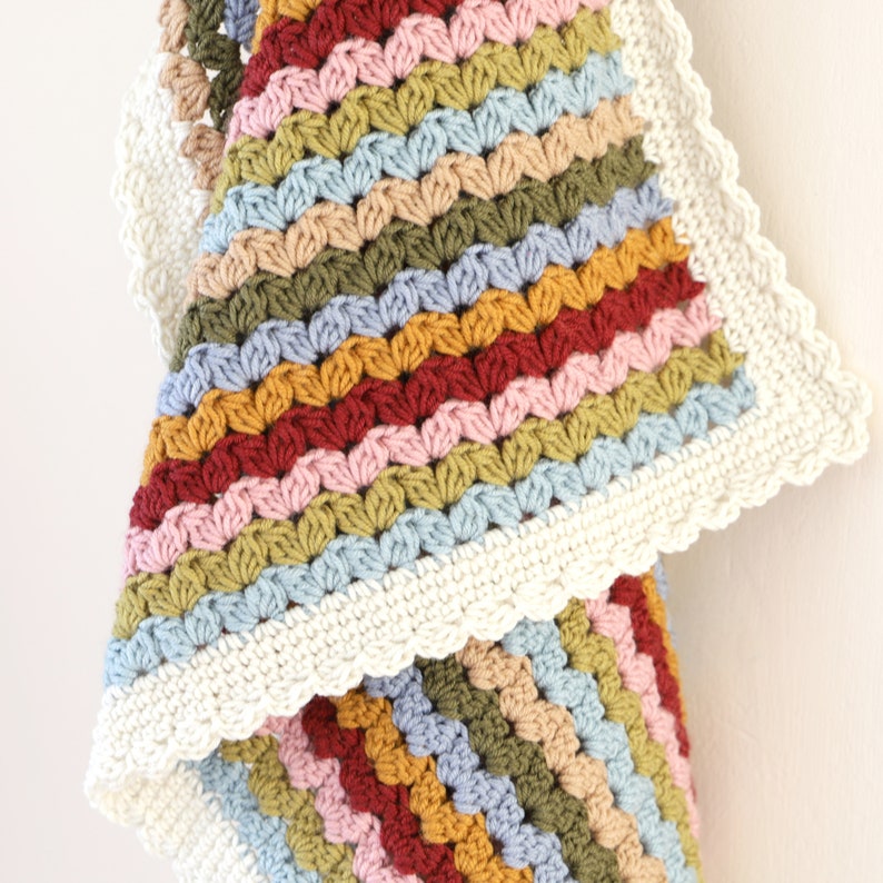 May include: A multi-colored crocheted baby blanket with a white crocheted border. The blanket is made with a variety of colors, including pink, red, yellow, green, and blue.
