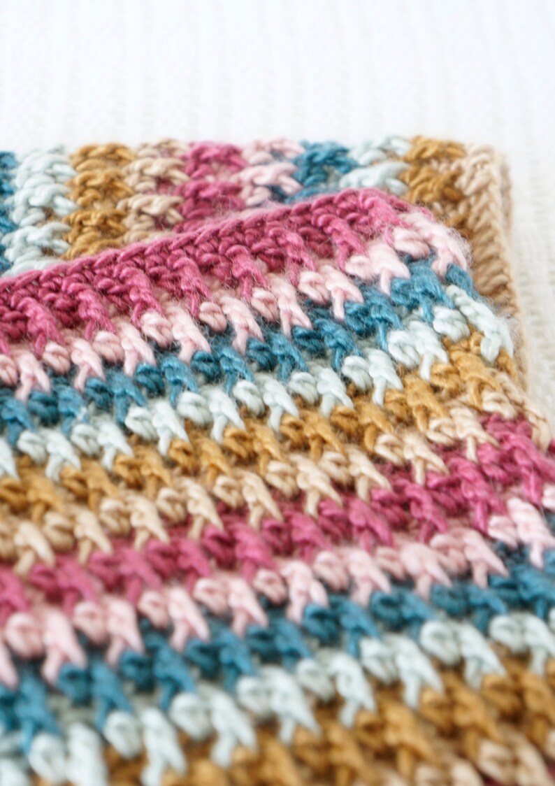 Puede incluir: Primer plano de una manta de ganchillo multicolor con rayas de color rosa, beige, azul claro y azul oscuro.