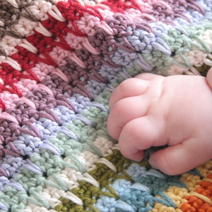 May include: A close-up of a baby's hand resting on a colorful crocheted blanket. The blanket is made with a variety of colors, including red, pink, purple, blue, green, and yellow.