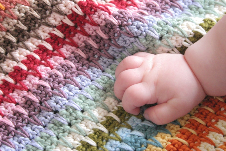 May include: A close-up of a baby's hand resting on a colorful crocheted blanket. The blanket is made with a variety of colors, including red, pink, purple, blue, green, and yellow.