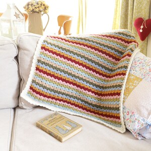 May include: A colorful striped crocheted throw blanket with a white border, draped over a white couch with a book titled "The Merry Animal Tales" by Marjorie Flack.
