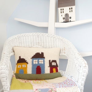 May include: A white wicker chair with a crocheted pillow featuring three colorful houses. The pillow is on top of a patchwork quilt with floral and geometric patterns. A small white house figurine sits on a white shelf in the background.