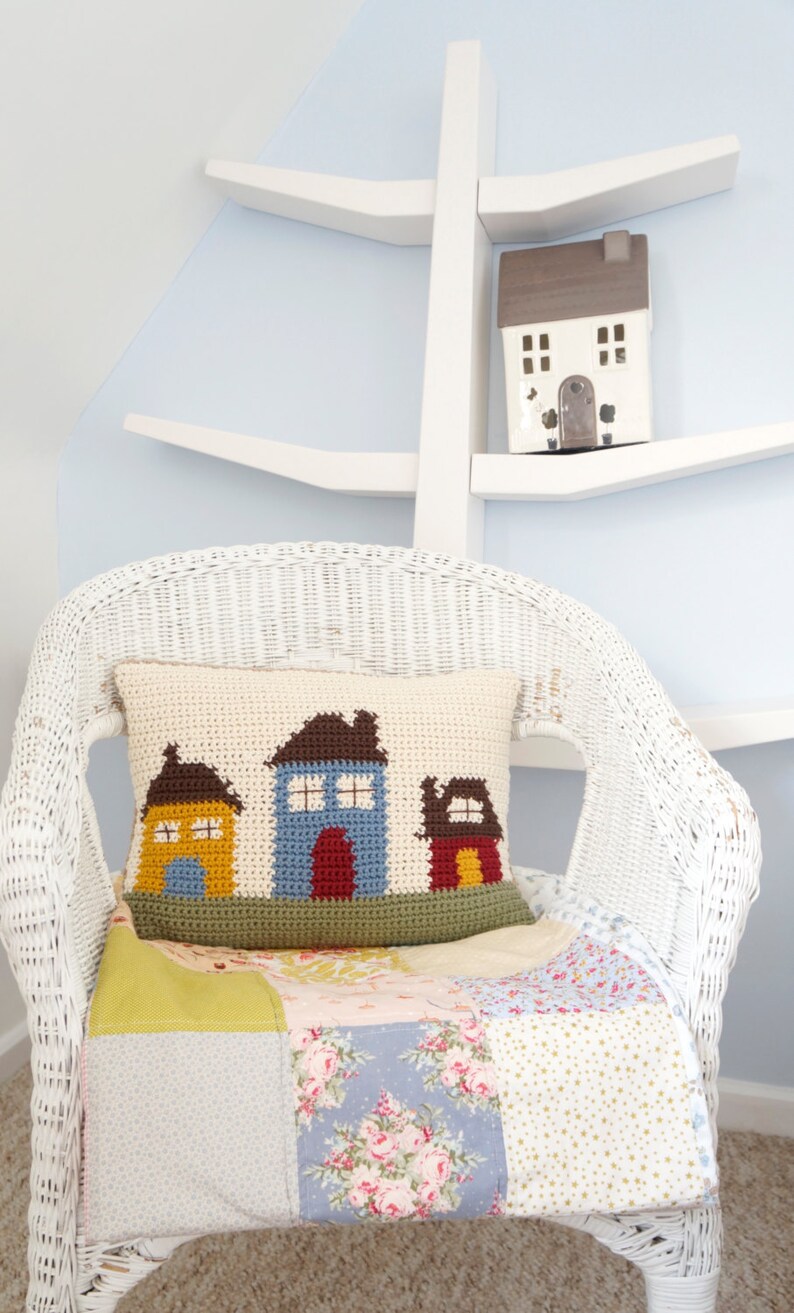 May include: A white wicker chair with a crocheted pillow featuring three colorful houses. The pillow is on top of a patchwork quilt with floral and geometric patterns. A small white house figurine sits on a white shelf in the background.