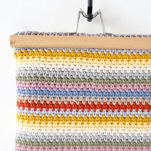 May include: A colorful crocheted wall hanging with a striped pattern in shades of yellow, orange, red, pink, blue, and green. The hanging is attached to a wooden dowel rod and a metal hanger.