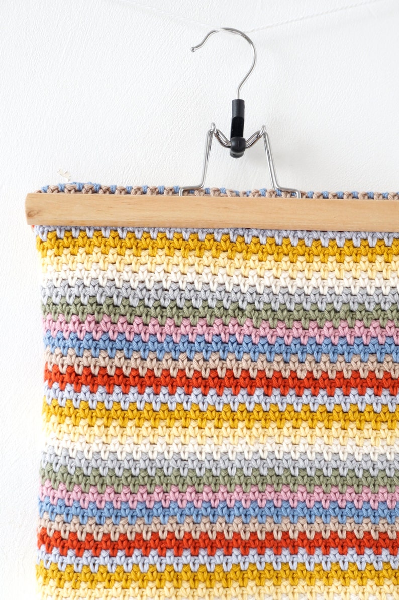 May include: A colorful crocheted wall hanging with a striped pattern in shades of yellow, orange, red, pink, blue, and green. The hanging is attached to a wooden dowel rod and a metal hanger.