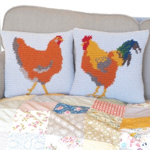 May include: Two crocheted throw pillows with a rooster and hen design. The pillows are on a couch with a patchwork quilt.