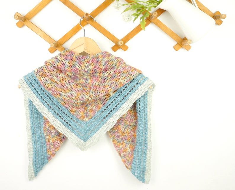 May include: A handmade crochet shawl in a triangular shape, featuring a hood. The shawl has a multicolored pattern with pink, yellow, and blue hues. The edges are trimmed with a light blue border and a white accent.
